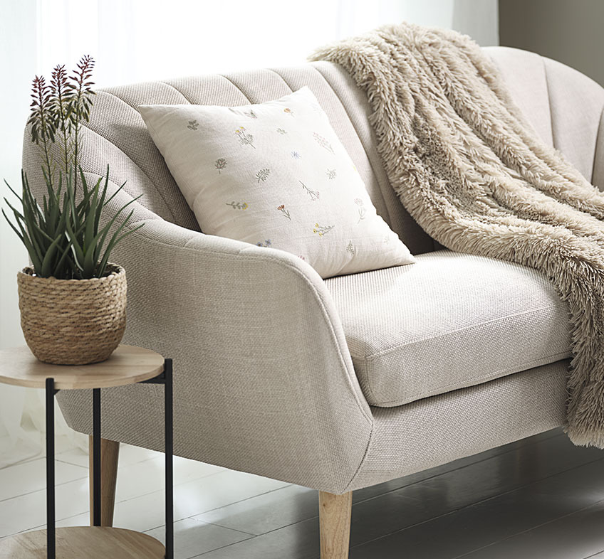 Pianta artificiale RASMUS in vaso STEFAN, cuscino AGERTIDSEL e coperta MYRFIOL Pianta artificiale di aloe vera con fiori, cuscino ricamato e lancio in pelliccia sintetica su divano beige