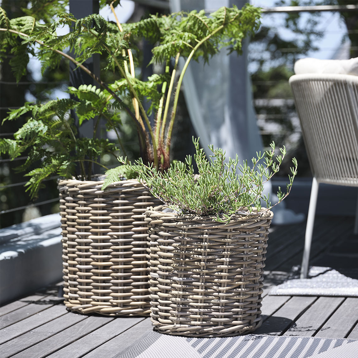 Due cestini porta-piante in rattan naturale non trattato posizionati su un balcone, con piante rigogliose che aggiungono un tocco di verde e freschezza allo spazio esterno