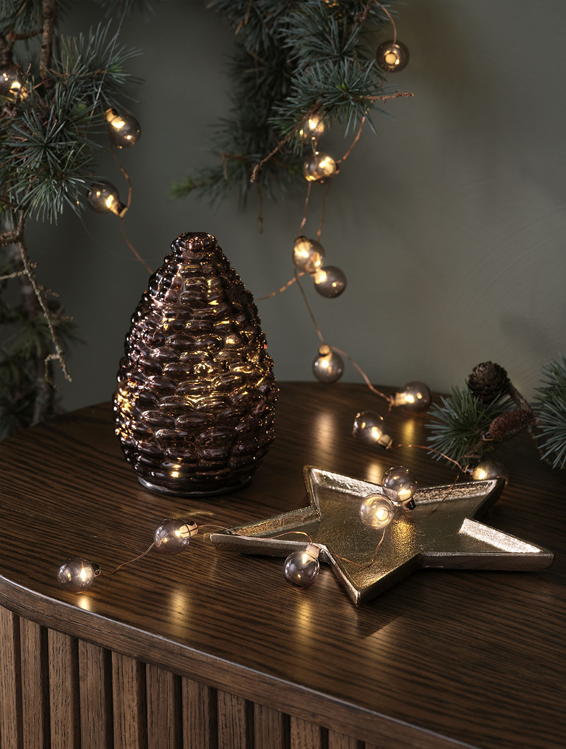 Pigna di Natale DELLING e catena di luci ALV Luci di Natale decorative, come una pigna di vetro e una catena di luci con sfere dorate, posizionate sulla parte superiore di una credenza.