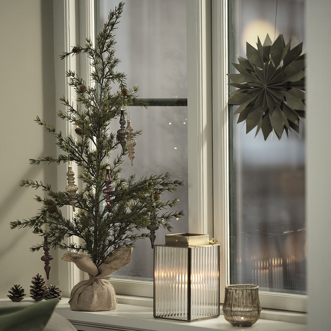 Albero FENRIR con decorazioni LUCIA, lanterna UVIT, portacandele SAFRAN e stella HRYM Portacandele, lanterna e albero artificiale posizionati su un davanzale